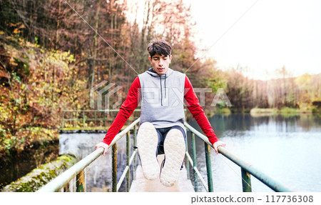 Sporty man doing l-sit outdoors, in park by lake. Morning workout for young man. 117736308