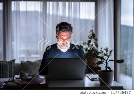 Handsome man working from home office at evening, using laptop. Screen glow illuminates man's face. Telecommuting and home office. Handsome man working from home office at evening, using laptop. Screen glow illuminates man's face. Telecommuting and home office. 117736427