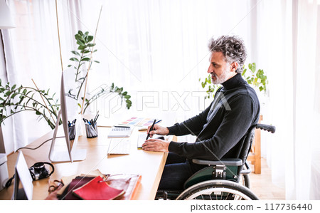 Handsome man in wheelchair working from home office, using graphics tablet and stylus. Telecommuting and home office. Handsome man in wheelchair working from home office, using graphics tablet and stylus. Telecommuting and home office. 117736440