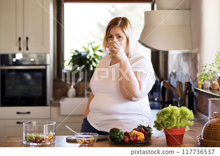 Overweight woman in the kitchen, drinking cup of fresh tap water. Choosing healthy diet, making vegetable salad. 117736537