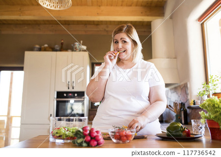 Overweight woman in the kitchen, making homemade lunch. Choosing healthy food, making and eating vegetable salad. 117736542