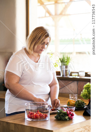 Overweight woman in the kitchen, making homemade lunch. Choosing healthy food, making and eating vegetable salad. 117736543