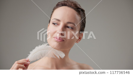 Beauty, health, cosmetics, anti-aging therapy and skin care concept - young beautiful brunette Caucasian woman holding a white fluffy feather, concept of tenderness, softness and lightness 117736546