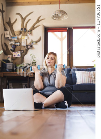 Online fitness training for overweight woman exercising at home with dumbbells, looking at laptop. Workout in living room. Online fitness training for overweight woman exercising at home with dumbbells, looking at laptop. Workout in living room. 117736551