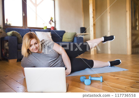 Online fitness training for overweight woman exercising on gym mat at home, looking at laptop. Workout in living room. 117736558