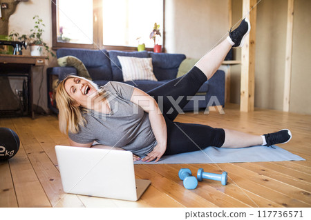 Online fitness training for overweight woman exercising on gym mat at home, looking at laptop. Workout in living room. 117736571
