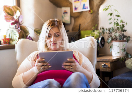 Portrait of beautiful young overweight woman at home, watching tv show on tablet, sitting in armchair. Portrait of beautiful young overweight woman at home, watching tv show on tablet, sitting in armchair. 117736574