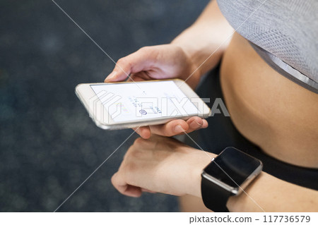 Young fitness girl checking her performance on smatphone and smartwatch after hard workout in gym. Young fitness girl checking her performance on smatphone and smartwatch after hard workout in gym. 117736579