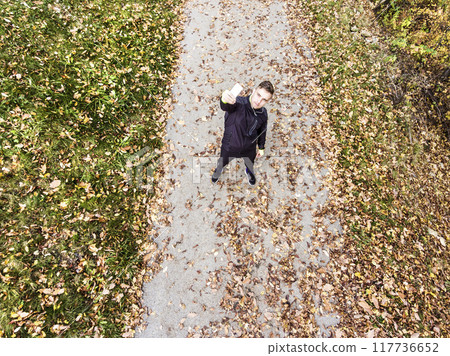 Aerial view of a runner standing autumn park on path with fallen leaves, holding smartphone and taking selfie. Morning running training. Aerial view of a runner standing autumn park on path with fallen leaves, holding smartphone and taking selfie. Morning running training. 117736652