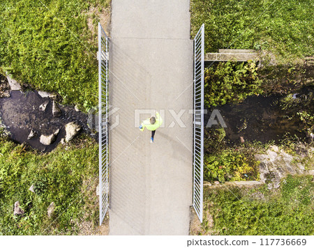 Aerial view of a runner running across bridge in park on a jogging path. Morning running training. Aerial view of a runner running across bridge in park on a jogging path. Morning running training. 117736669