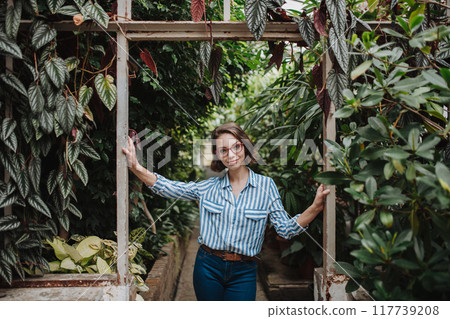 Small greenhouse business. Businesswoman selling flowers and houseplants, standing in greenhouse, looking at camera, smiling. Small greenhouse business. Businesswoman selling flowers and houseplants, standing in greenhouse, looking at camera, smiling. 117739208