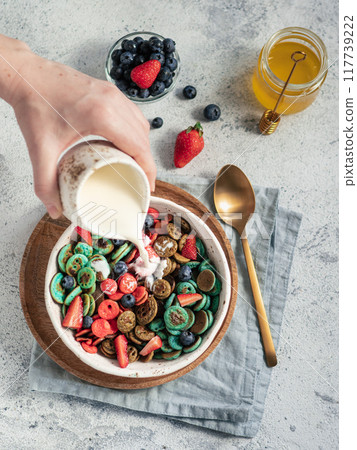 Trendy food - pancake cereal. Milk pouring on heap of multicolor mini cereal pancakes in bowl. Tiny cereal pancakes with berries in craft plate over gray cement background. Vertical. Top view,flat lay Trendy food - pancake cereal. Milk pouring on heap of multicolor mini cereal pancakes in bowl. Tiny cereal pancakes with berries in craft plate over gray cement background. Vertical. Top view,flat lay 117739222
