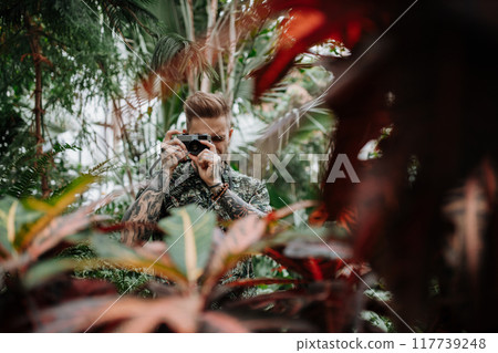 Handsome man with tattoos standing in botanical garden, in the middle of lush green foliage, holding camera and taking photography. Handsome man with tattoos standing in botanical garden, in the middle of lush green foliage, holding camera and taking photography. 117739248