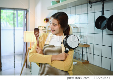 Portrait of young woman holding wooden spatula and pot standing in a kitchen. Domestic life and cooking concept 117740635