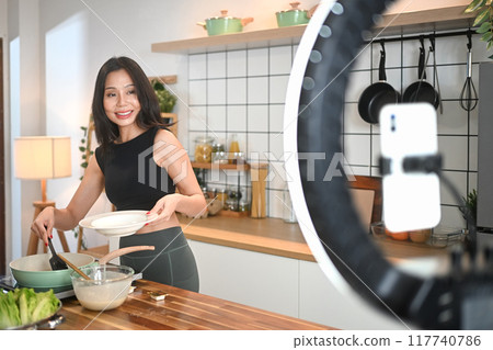 Attractive sporty woman recording a video for a cooking tutorial or vlog while preparing a healthy meal in a kitchen Attractive sporty woman recording a video for a cooking tutorial or vlog while preparing a healthy meal in a kitchen 117740786