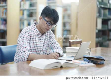 Young Asian male sitting inside a library alone doing research. Man working on a project. Young man doing research for a case. Young Asian male sitting inside a library alone doing research. Man working on a project. Young man doing research for a case. 117742311