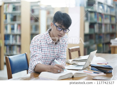 Young Asian male sitting inside a library alone doing research. Man working on a project. Young man doing research for a case. 117742327