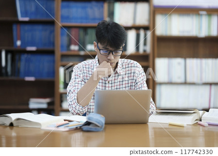 Young Asian male sitting inside a library alone doing research. Man working on a project. Young man doing research for a case. Young Asian male sitting inside a library alone doing research. Man working on a project. Young man doing research for a case. 117742330