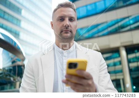 Focused young corporate worker investor businessman walking and using mobile phone outdoors near a business centre. Focused young corporate worker investor businessman walking and using mobile phone outdoors near a business centre. 117743283