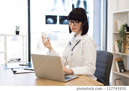 Female doctor working on laptop and holding medicine bottle 117743830