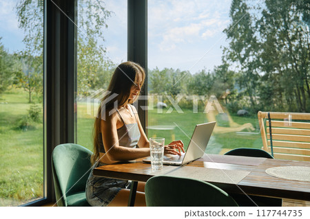 Woman with smooth long hair typing on laptop with lush green landscape outside Woman with smooth long hair typing on laptop with lush green landscape outside 117744735