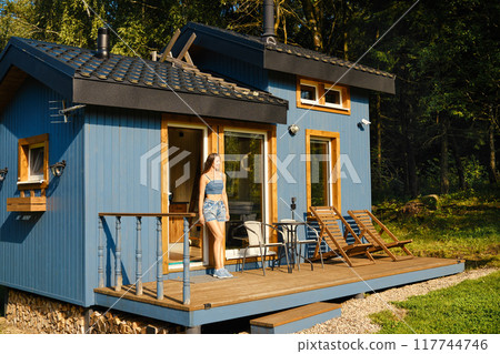 Woman leaning against the doorway of cozy tiny house surrounded by trees in a sunny forest clearing 117744746