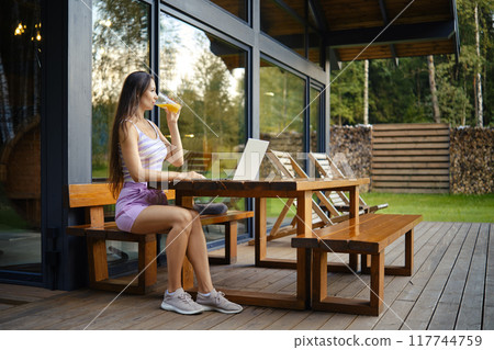 Woman enjoys a drink while using laptop on patio of a modern cabin surrounded by nature 117744759