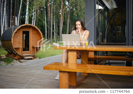 Woman watching movie on a laptop while sitting on a wooden bench on the terrace of a country house Woman watching movie on a laptop while sitting on a wooden bench on the terrace of a country house 117744760