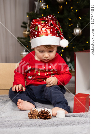A one-year-old kid in a Santa hat opens gifts near the Christmas tree A one-year-old kid in a Santa hat opens gifts near the Christmas tree 117744813