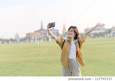 Traveler asian woman in her 30s making a livestream or selfie on smartphone while explores Wat Phra Kaew emerald Buddha. Share the wonders of Thai heritage through her journey. Traveler asian woman in her 30s making a livestream or selfie on smartphone while explores Wat Phra Kaew emerald Buddha. Share the wonders of Thai heritage through her journey. 117745317
