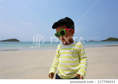 A boy about two years old wearing a long-sleeved shirt and wear sunglasses, walking on the beach smiling happily. 117746033