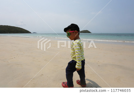 A boy about two years old wearing a long-sleeved shirt and wear sunglasses, walking on the beach smiling happily. 117746035