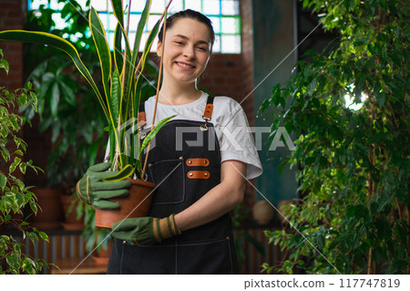 Running of own business. Young woman florist holding plant in pot wearing apron in botanical store. Happy small business owner working at flower shop smiling surrounded by plants. Small business Running of own business. Young woman florist holding plant in pot wearing apron in botanical store. Happy small business owner working at flower shop smiling surrounded by plants. Small business 117747819
