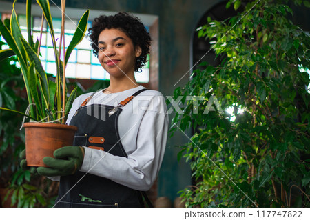 Running of own business. African woman florist holding plant in pot wearing apron in botanical store. Happy small business owner working at flower shop smiling surrounded by plants. Small business Running of own business. African woman florist holding plant in pot wearing apron in botanical store. Happy small business owner working at flower shop smiling surrounded by plants. Small business 117747822