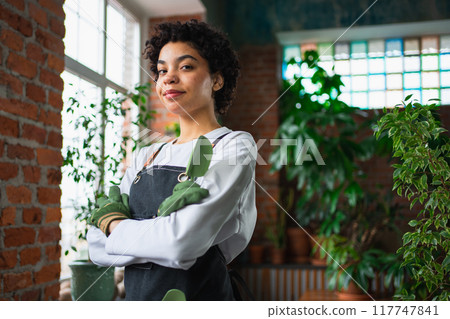 Running of own business. African woman florist wearing apron in botanical store with green plants. Happy small business owner working at flower shop smiling surrounded by plants Small business Running of own business. African woman florist wearing apron in botanical store with green plants. Happy small business owner working at flower shop smiling surrounded by plants Small business 117747841