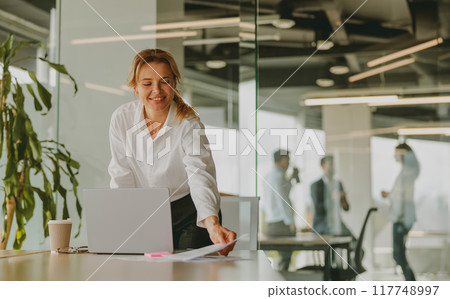 A Smiling Businesswoman is Engaged in Working on Her Laptop Inside a Modern Office Environment A Smiling Businesswoman is Engaged in Working on Her Laptop Inside a Modern Office Environment 117748997