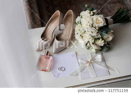Bride's wedding essentials: heels, bouquet, rings, perfume, and invitation on a white table 117751139
