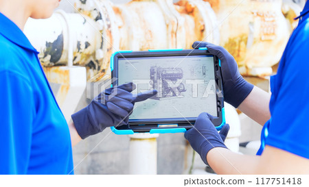 A worker inspecting pipes while looking at a tablet A worker inspecting pipes while looking at a tablet 117751418