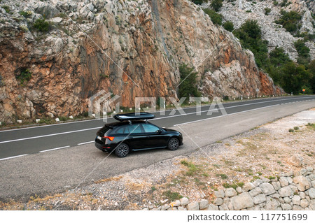 A black car parked near a rocky landscape along a winding mountain road.  117751699