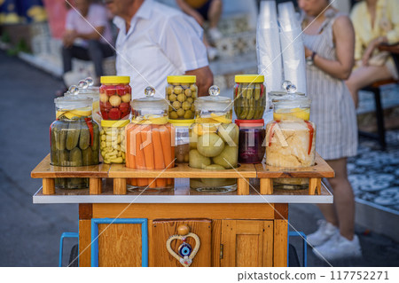 Colorful jars of pickled vegetables displayed on a wooden cart at a bustling market Colorful jars of pickled vegetables displayed on a wooden cart at a bustling market 117752271
