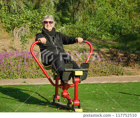 Older Woman in Sunglasses Engages in Morning Workout on Rowing Machine at Outdoor Park Gym, Promoting Active and Healthy Living for All Ages. Older Woman in Sunglasses Engages in Morning Workout on Rowing Machine at Outdoor Park Gym, Promoting Active and Healthy Living for All Ages. 117752510