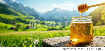 Honey is pouring from a wooden honey dipper into a glass jar, close-up, sunny rural landscape with flower meadows and hill, copy space 117754383