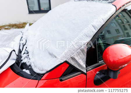 Close-up detail red car with snow-covered windshield protected by reflective cover mat, parked outside residential building during winter. Windscreen window ice frost protection 117755275