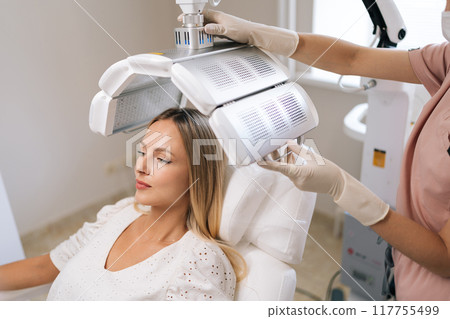 High-angle view of pretty blonde female having LED light hair photodynamic therapy treatment at modern medical clinic. Cosmetician specialist adjusting innovation device for PDT therapy procedure High-angle view of pretty blonde female having LED light hair photodynamic therapy treatment at modern medical clinic. Cosmetician specialist adjusting innovation device for PDT therapy procedure 117755499