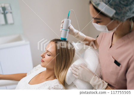 Closeup of trichologist in gloves and mask performing hair treatment procedure using spectrum phototherapy device. Blonde female patient having hair light therapy with modern innovation equipment. Closeup of trichologist in gloves and mask performing hair treatment procedure using spectrum phototherapy device. Blonde female patient having hair light therapy with modern innovation equipment. 117755505