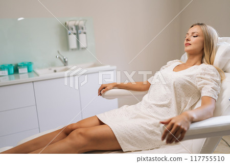 Pensive blonde young woman client in white procedure uniform lying on cosmetology chair waiting beautician specialist in modern beauty salon. Concept of cosmetology, spa, beauty treatment. Pensive blonde young woman client in white procedure uniform lying on cosmetology chair waiting beautician specialist in modern beauty salon. Concept of cosmetology, spa, beauty treatment. 117755510