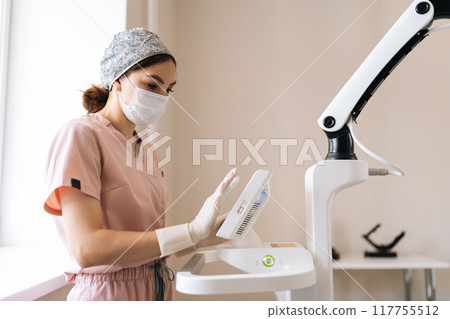 Focused female doctor in protective mask and gloves setting modern device in medical clinic. Side view of beautician specialist adjusting innovation device for skin rejuvenation at aesthetic clinic. Focused female doctor in protective mask and gloves setting modern device in medical clinic. Side view of beautician specialist adjusting innovation device for skin rejuvenation at aesthetic clinic. 117755512