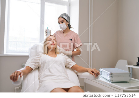 Portrait of trichologist in gloves and mask performing hair treatment procedure using spectrum phototherapy device. Blonde female patient having hair light therapy with modern innovation equipment. 117755514