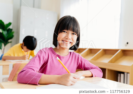 Enthusiastic young learner engages with camera during writing task. Smiling Asian preschooler showcases confidence and curiosity in classroom 117756329