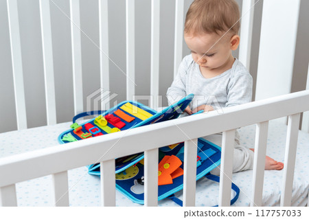Baby playing with montessori busy book sitting in crib. Concept of smart books and modern toys Baby playing with montessori busy book sitting in crib. Concept of smart books and modern toys 117757033